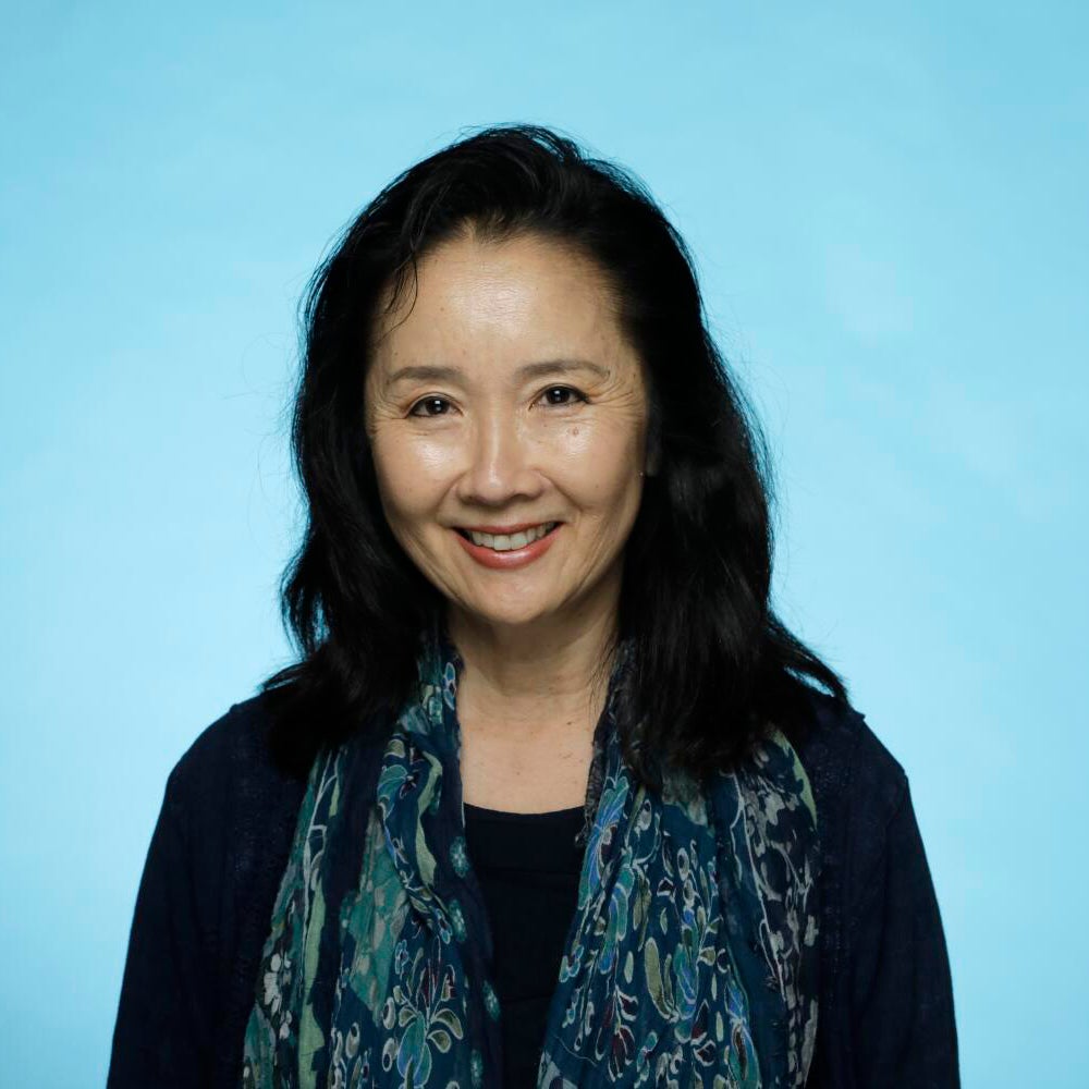 Teresa Watanabe smiles at the camera against a light blue background, wearing a dark top and patterned scarf.